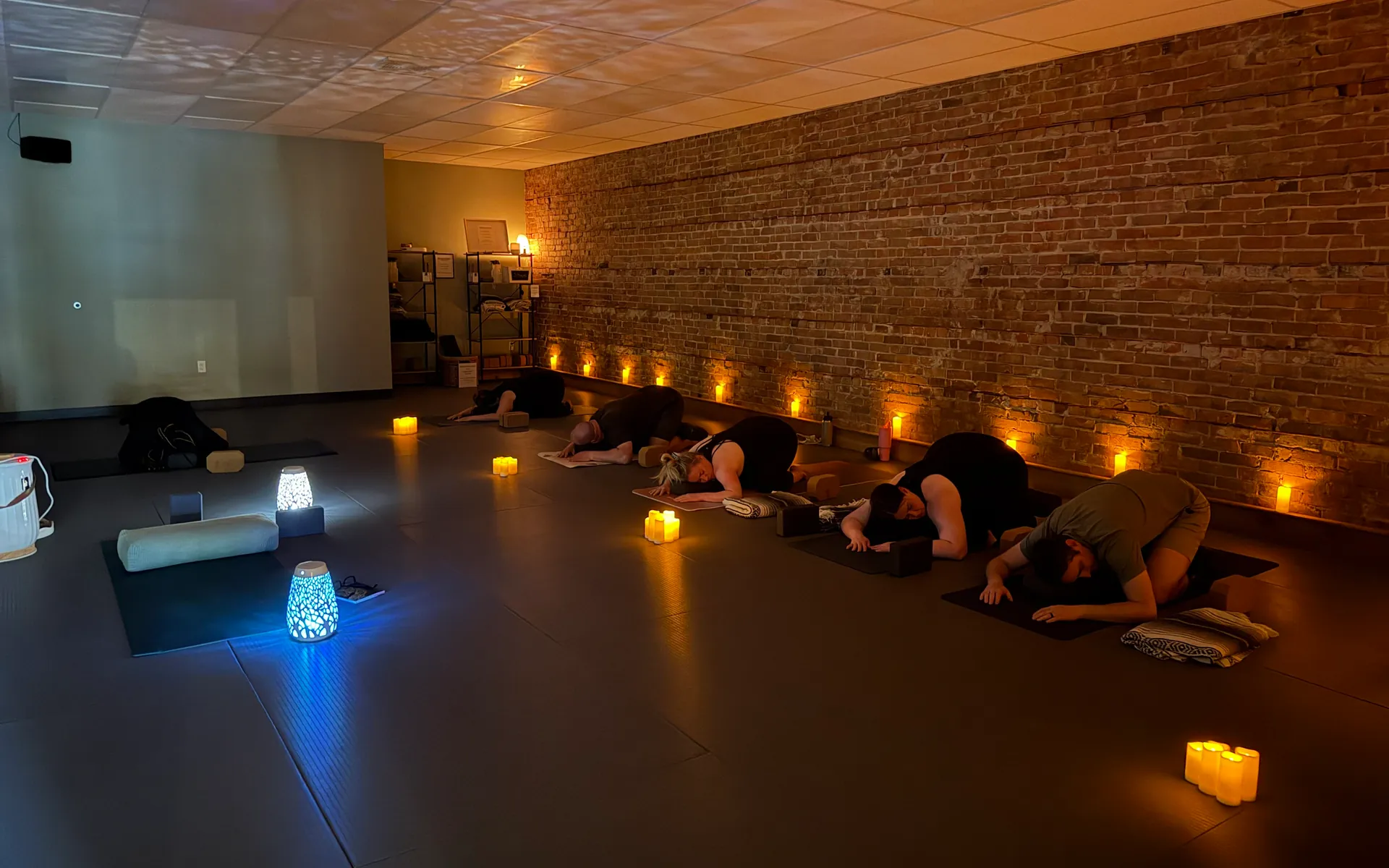 A candlelit class resting in child’s pose along the studio’s brick wall